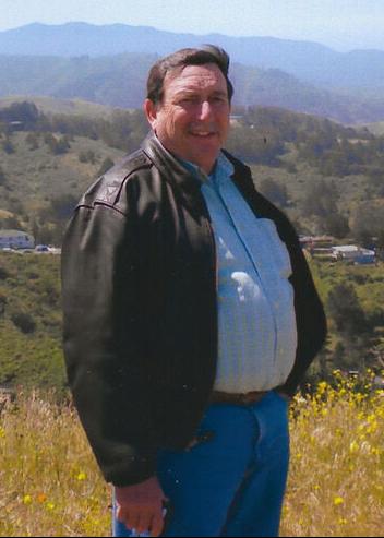 Ezio Francis Nurisio stands on a hillside on a sunny day 
wearing a leather jacket and looking at the camera